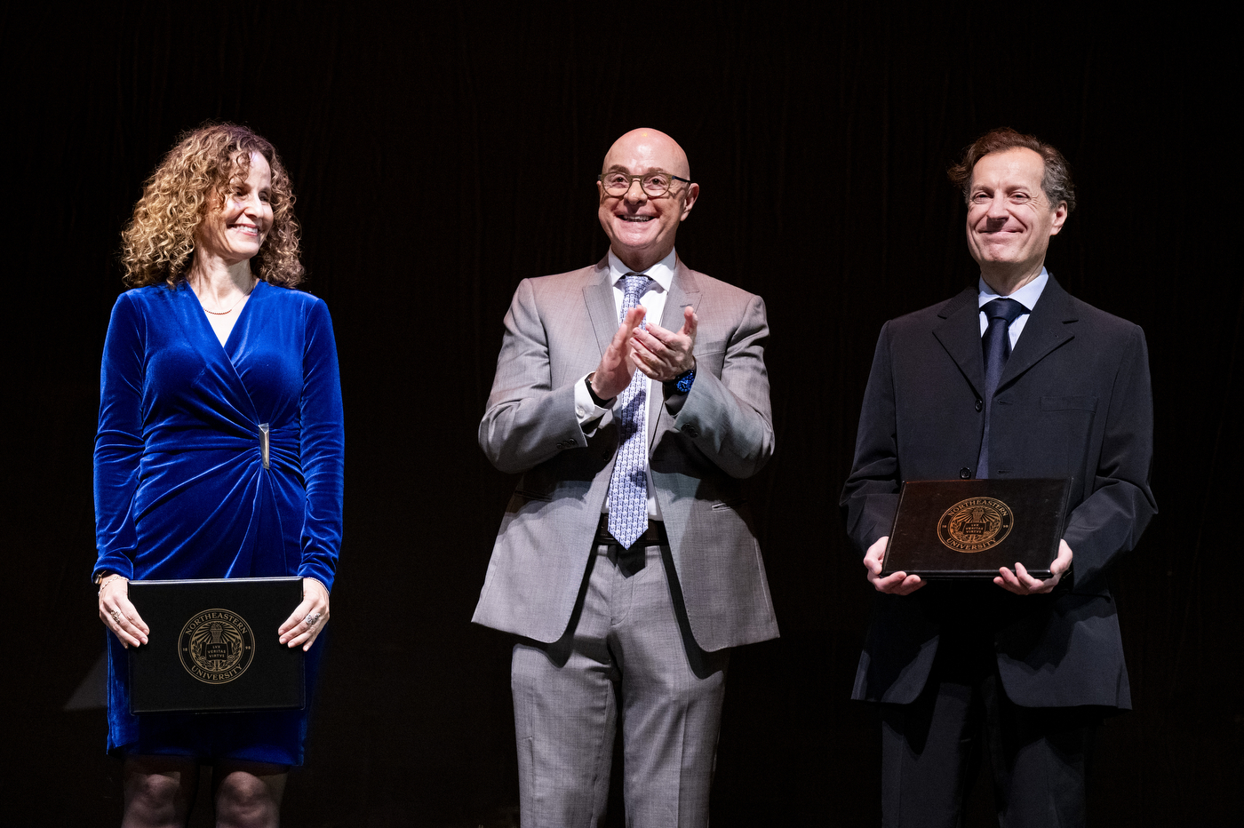 President Aoun stands on stage wearing a grey suit with two people on either side of him at the Academic Honors Convocation ceremony.