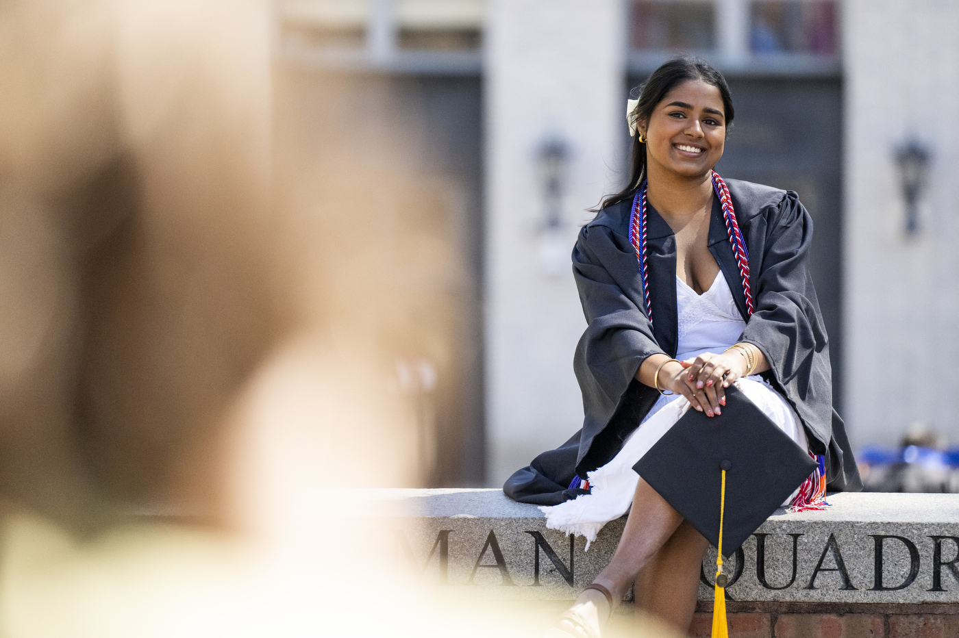 A student in regalia sits on a wall engraved with Northeastern University and poses for a photo.