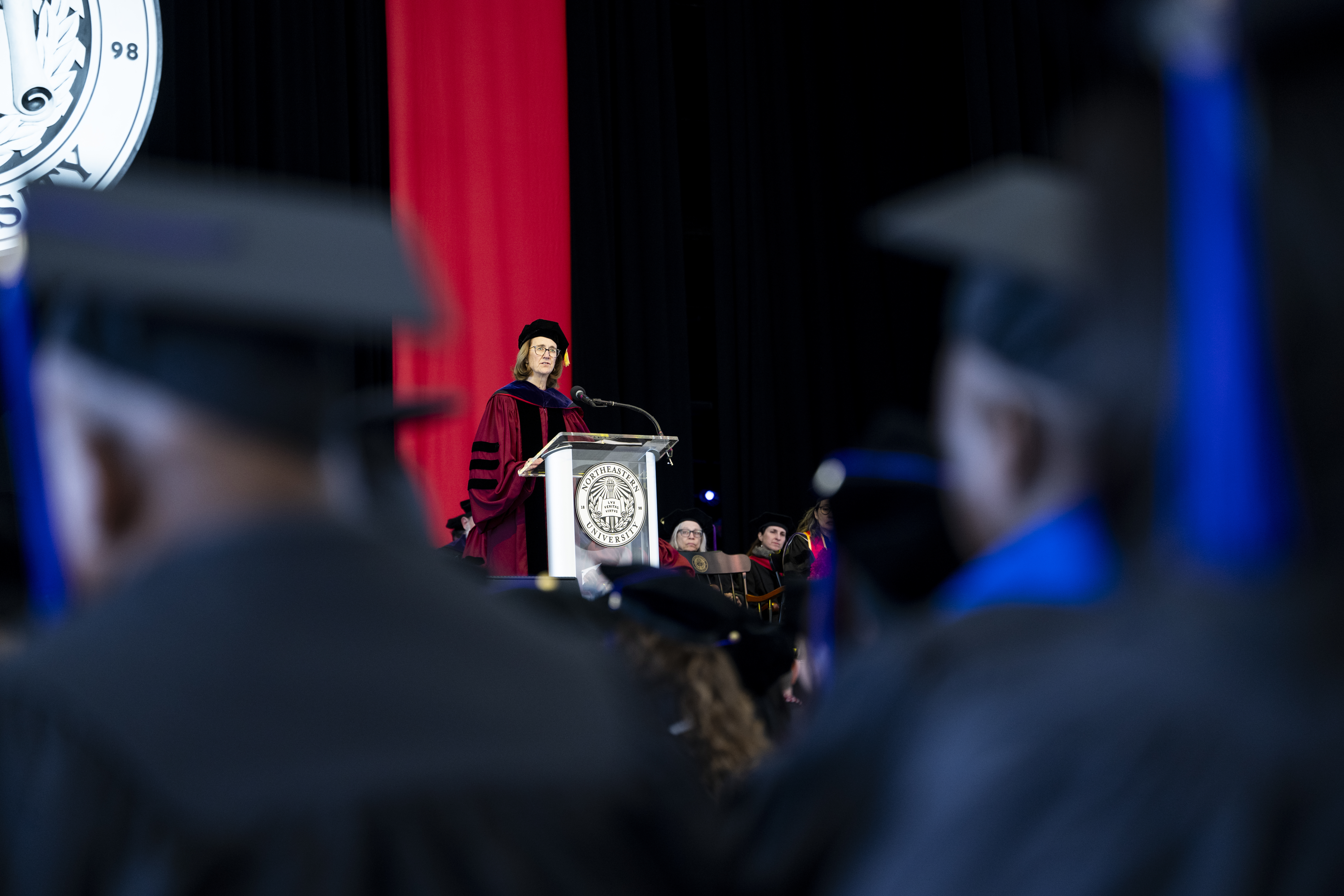 A wide shot shows Lois Cornell at a podium on stage, viewed past blurred graduates in caps and gowns in the foreground.