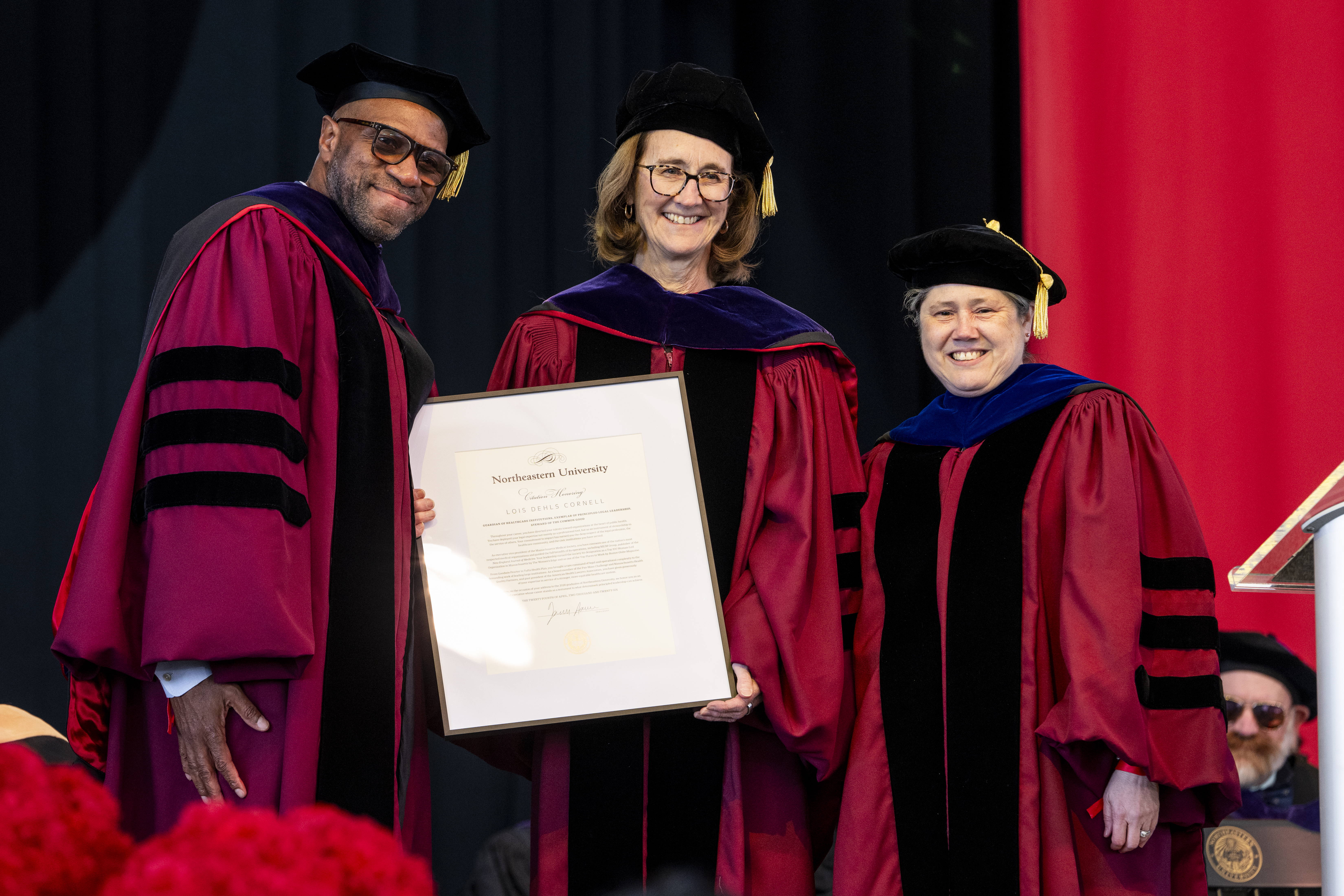 Three faculty members in red academic robes and black caps smile while holding a framed certificate on stage during a commencement ceremony.