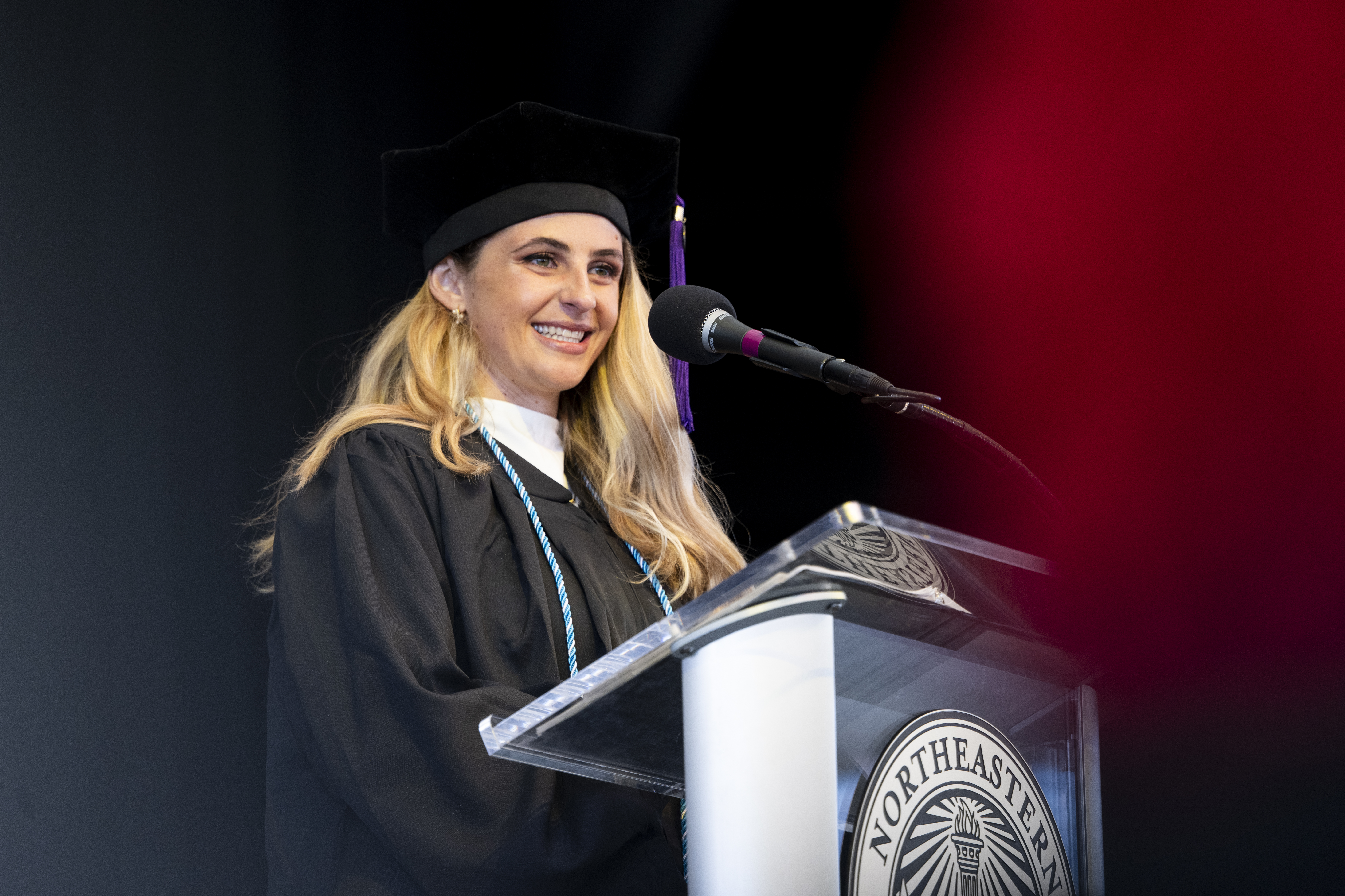 A smiling woman in a black graduation cap and gown stands at a podium speaking into a microphone, with a dark stage backdrop and a blurred red light along the edge.