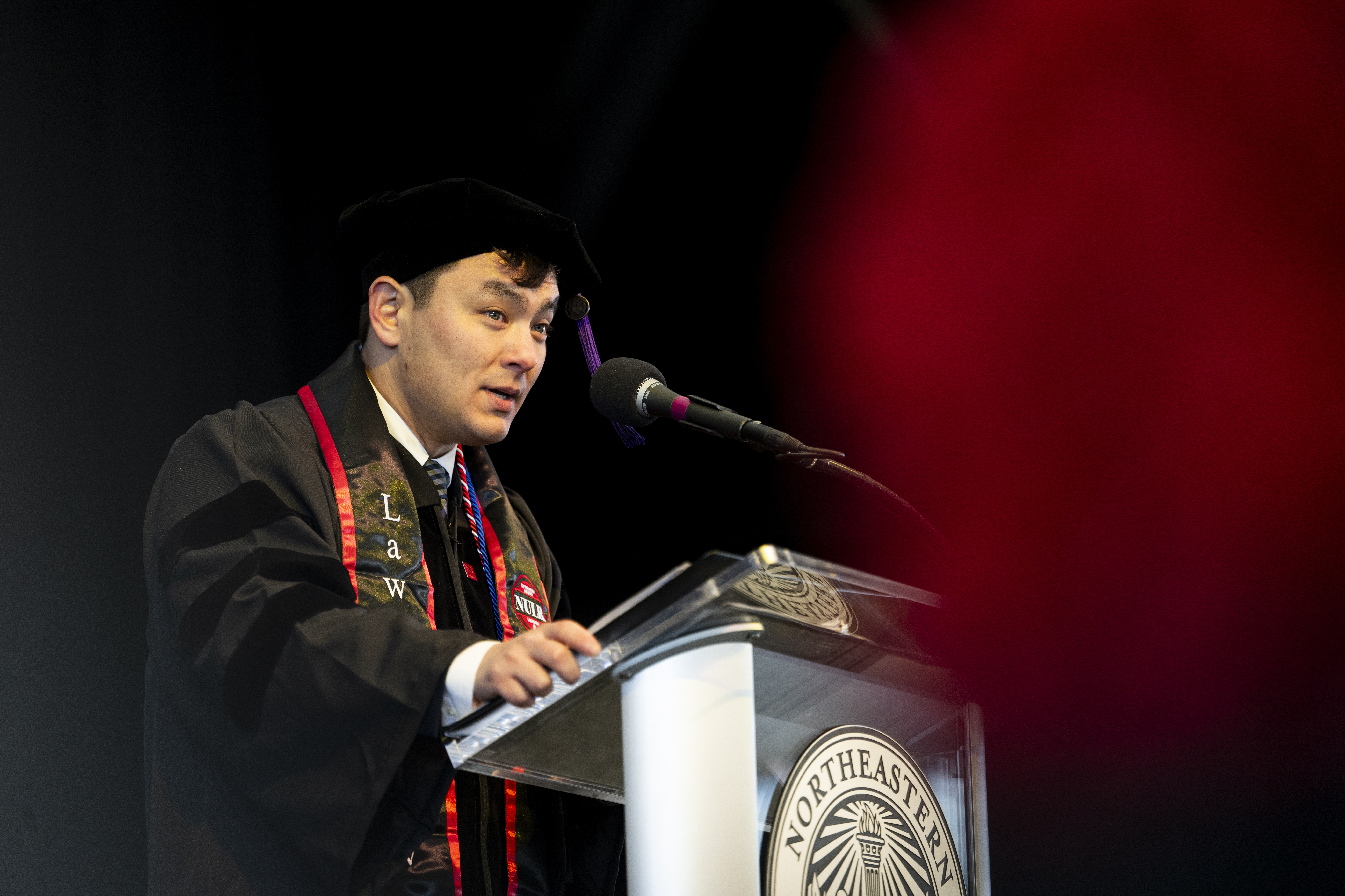 A graduate wearing a decorated stole and black cap and gown delivers a speech at a podium, speaking into a microphone with a dark background and red blur in the foreground.