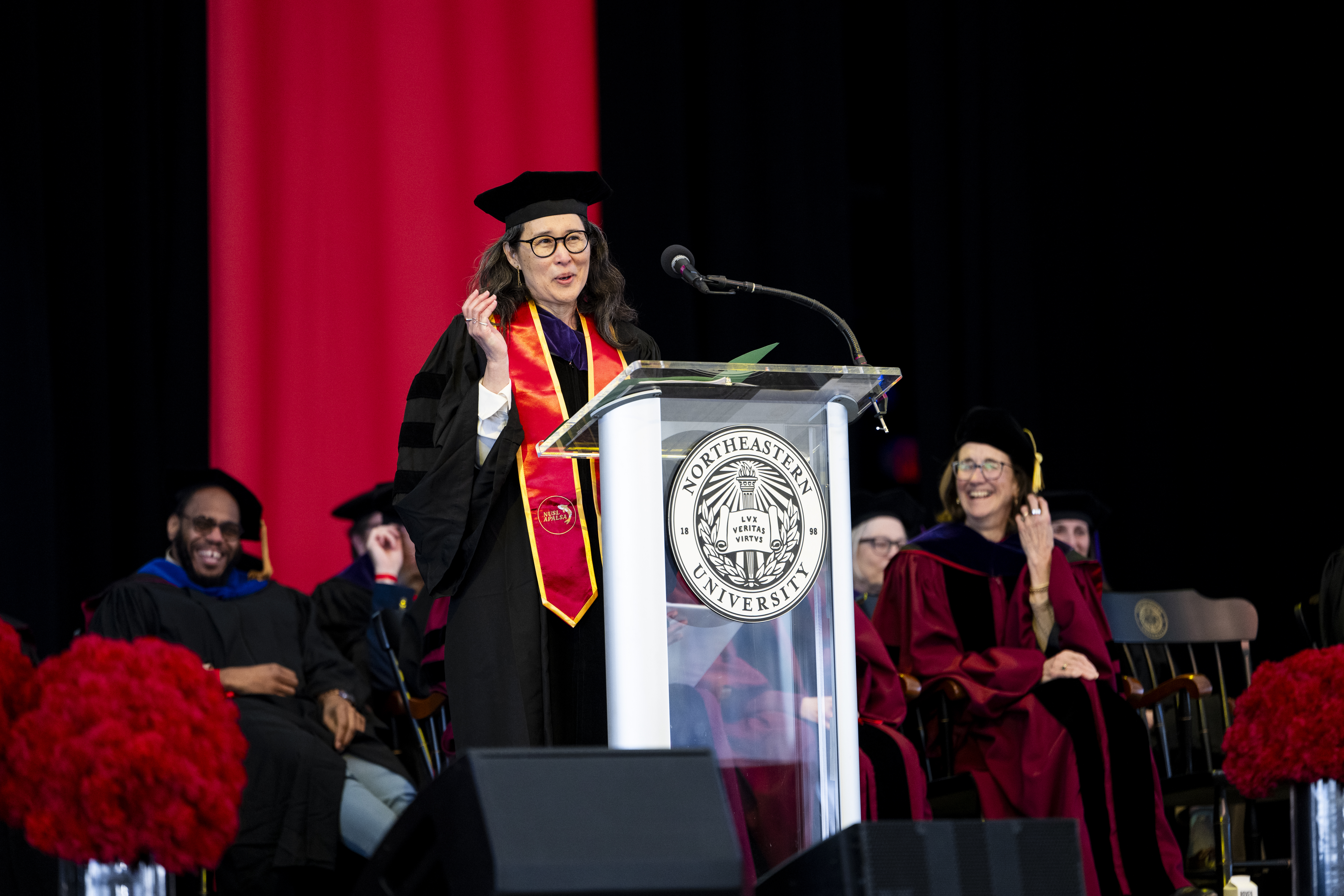 A commencement speaker in academic regalia stands at a podium with a Northeastern University seal, gesturing while speaking against a red stage backdrop as seated faculty smile behind her.