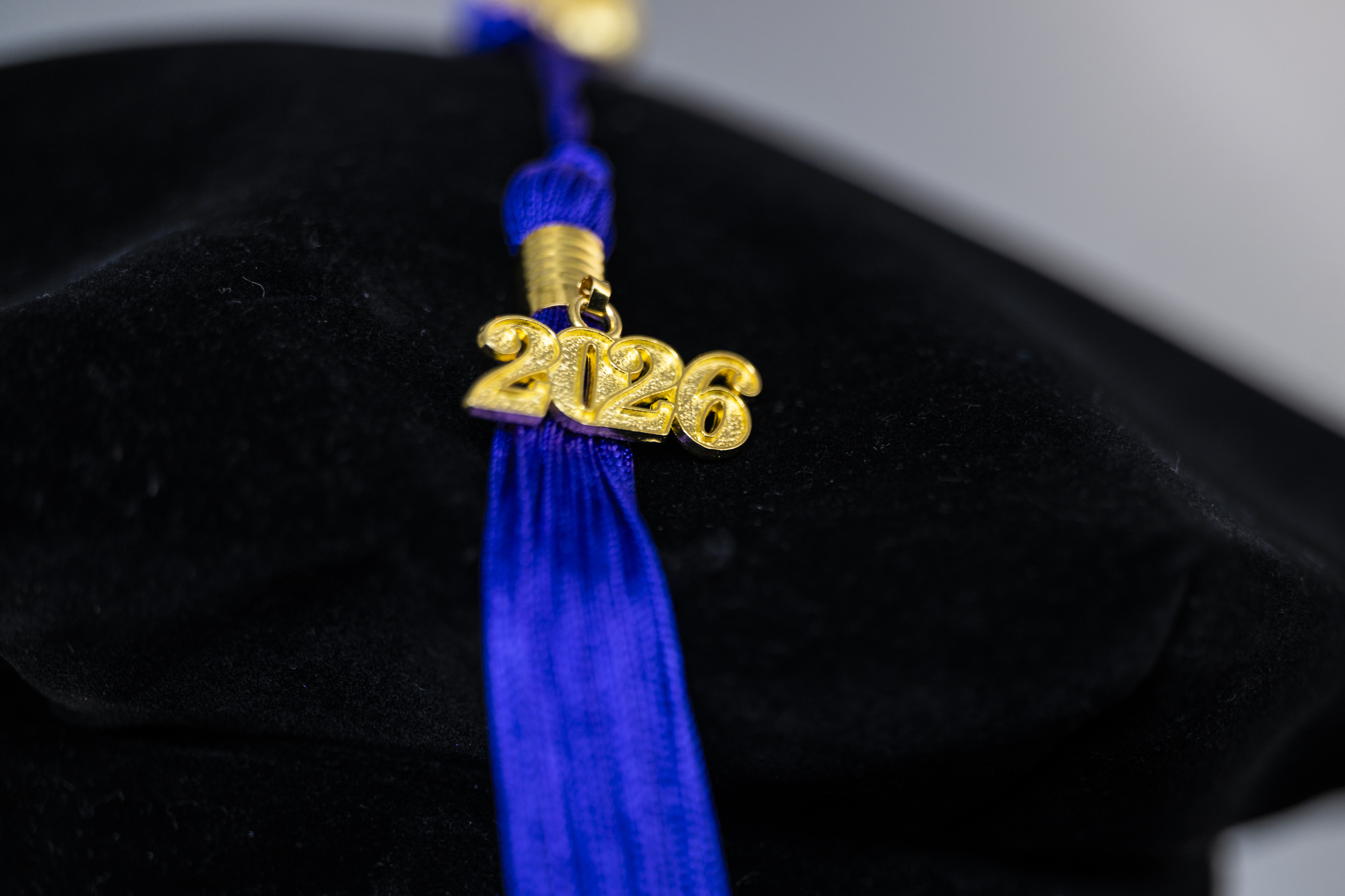 Close-up of a blue graduation tassel hanging from a cap, featuring a gold “2026” charm in sharp focus against a blurred dark background.