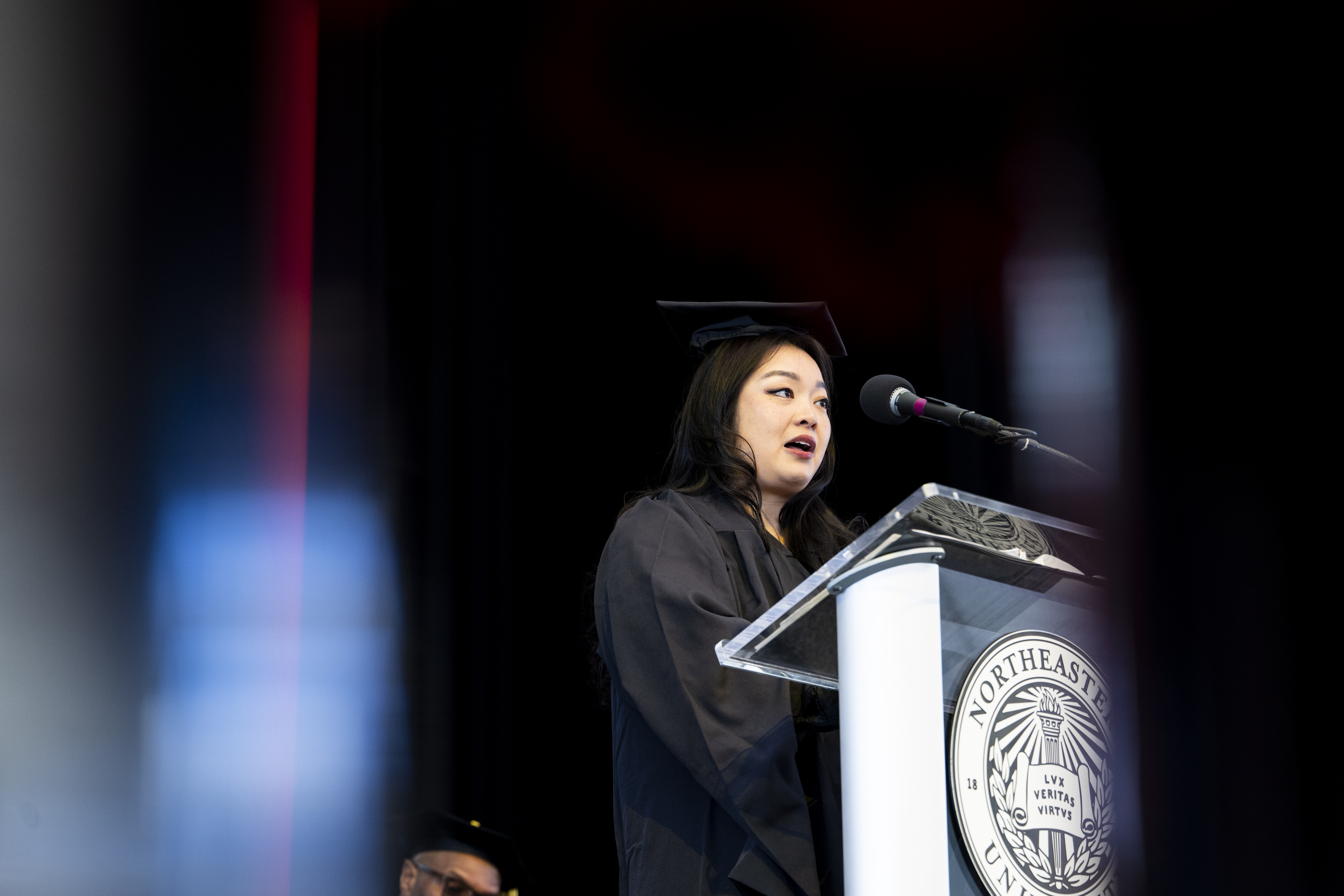 A graduate in a black cap and gown speaks at a podium on stage, facing a microphone, with a dark background and soft colored light streaks in the foreground.
