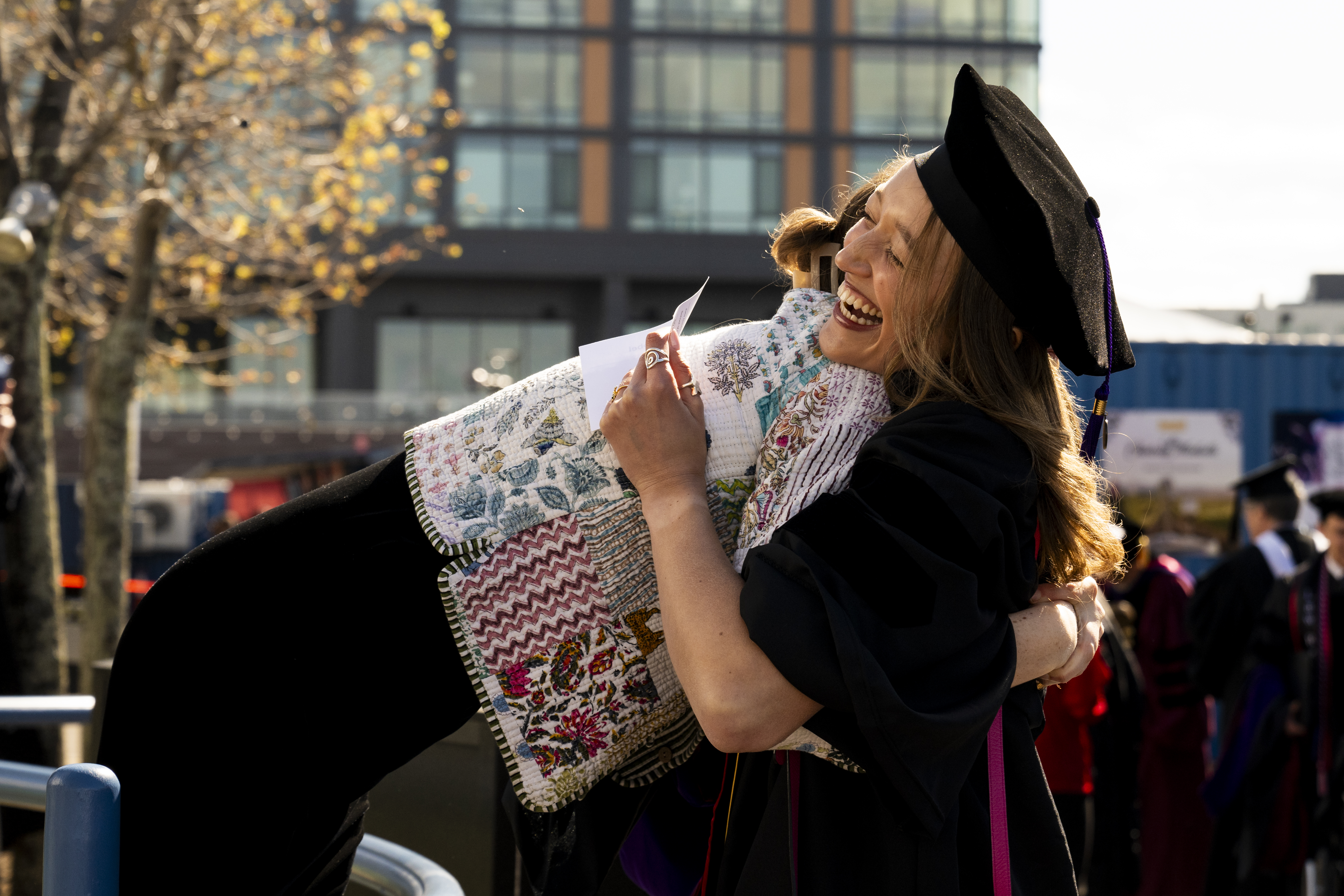Two graduates in black caps and gowns embrace outdoors, smiling and holding a patterned blanket, with campus buildings and trees in the background.