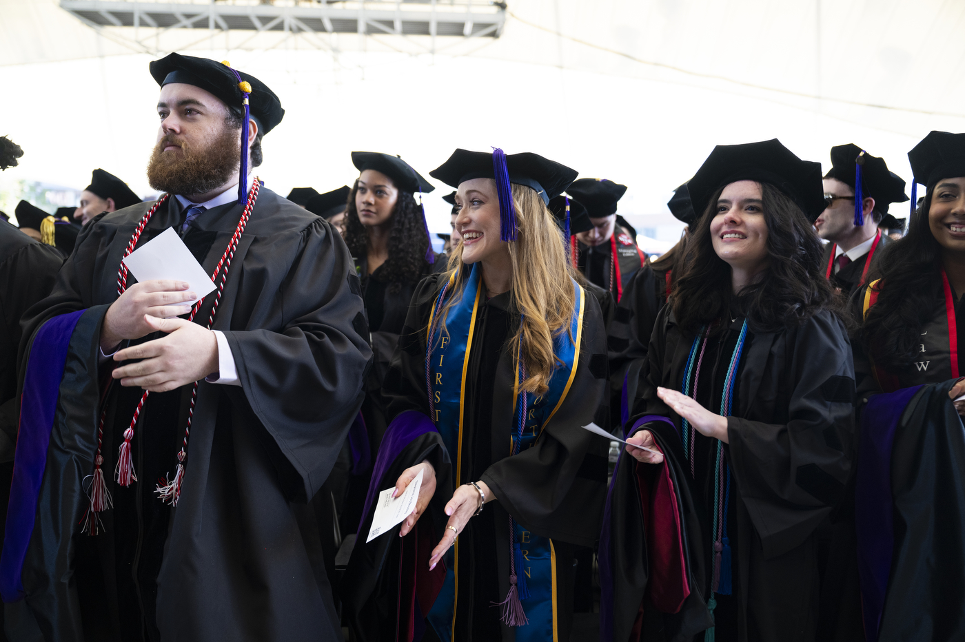 A group of graduates in black caps and gowns stand together indoors, smiling and chatting, some holding programs and wearing honor cords and stoles.