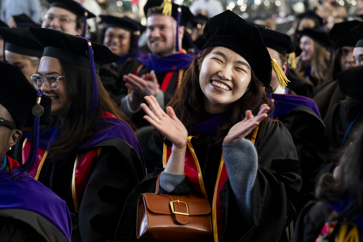 A group of graduates seated at commencement clap and smile, with one woman in the foreground beaming and applauding while wearing a black cap and gown.