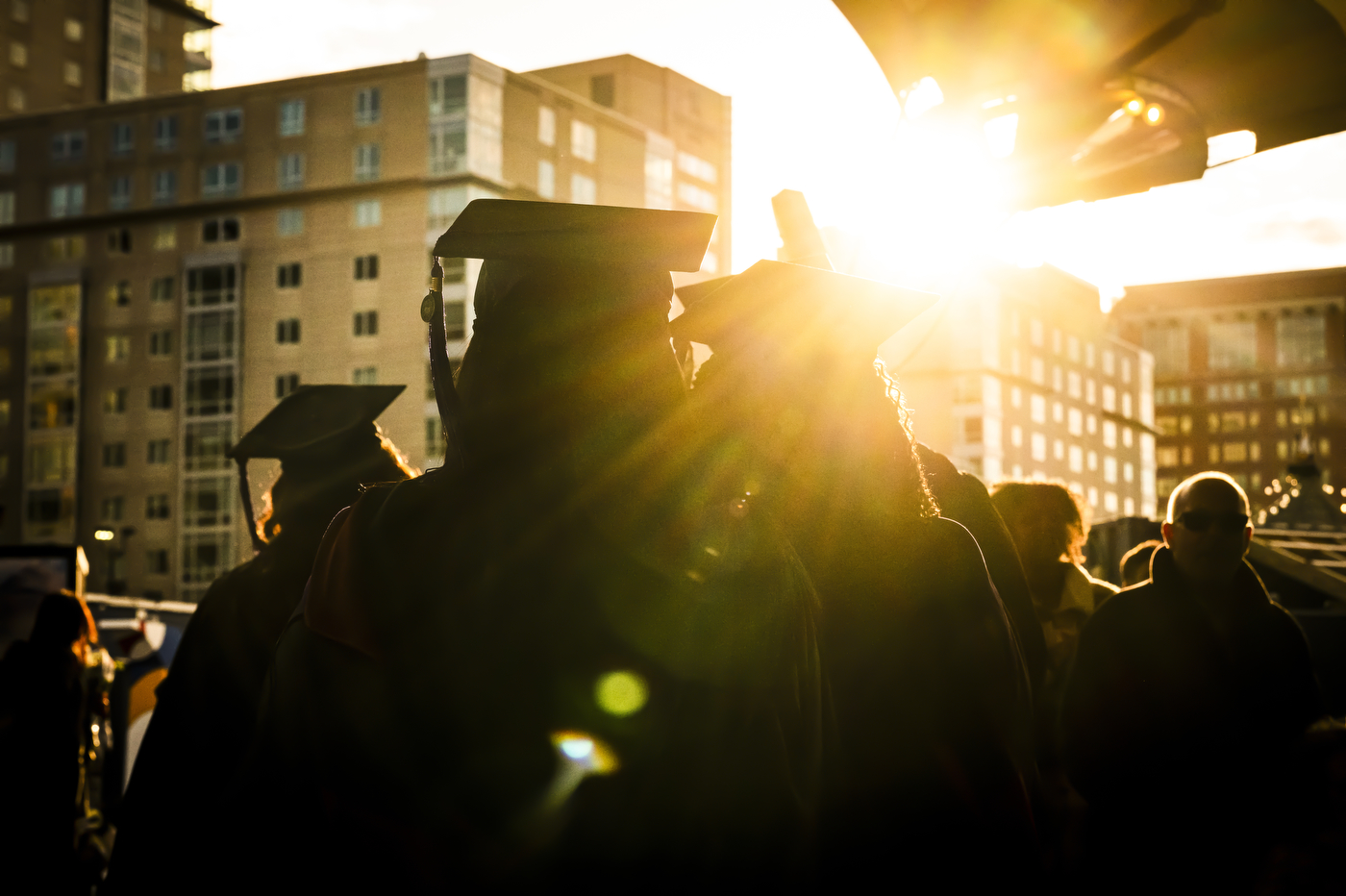 Silhouetted graduates in caps and gowns stand outdoors at sunset, with bright sunlight flaring between buildings in the background.