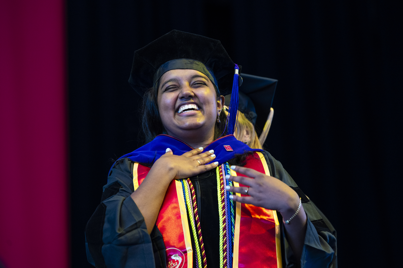 A graduate in a cap and gown smiles broadly on stage, placing a hand on their chest while wearing colorful honor cords and a red stole.