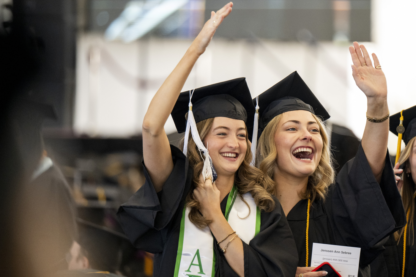 Two graduates in caps and gowns wave to the crowd.