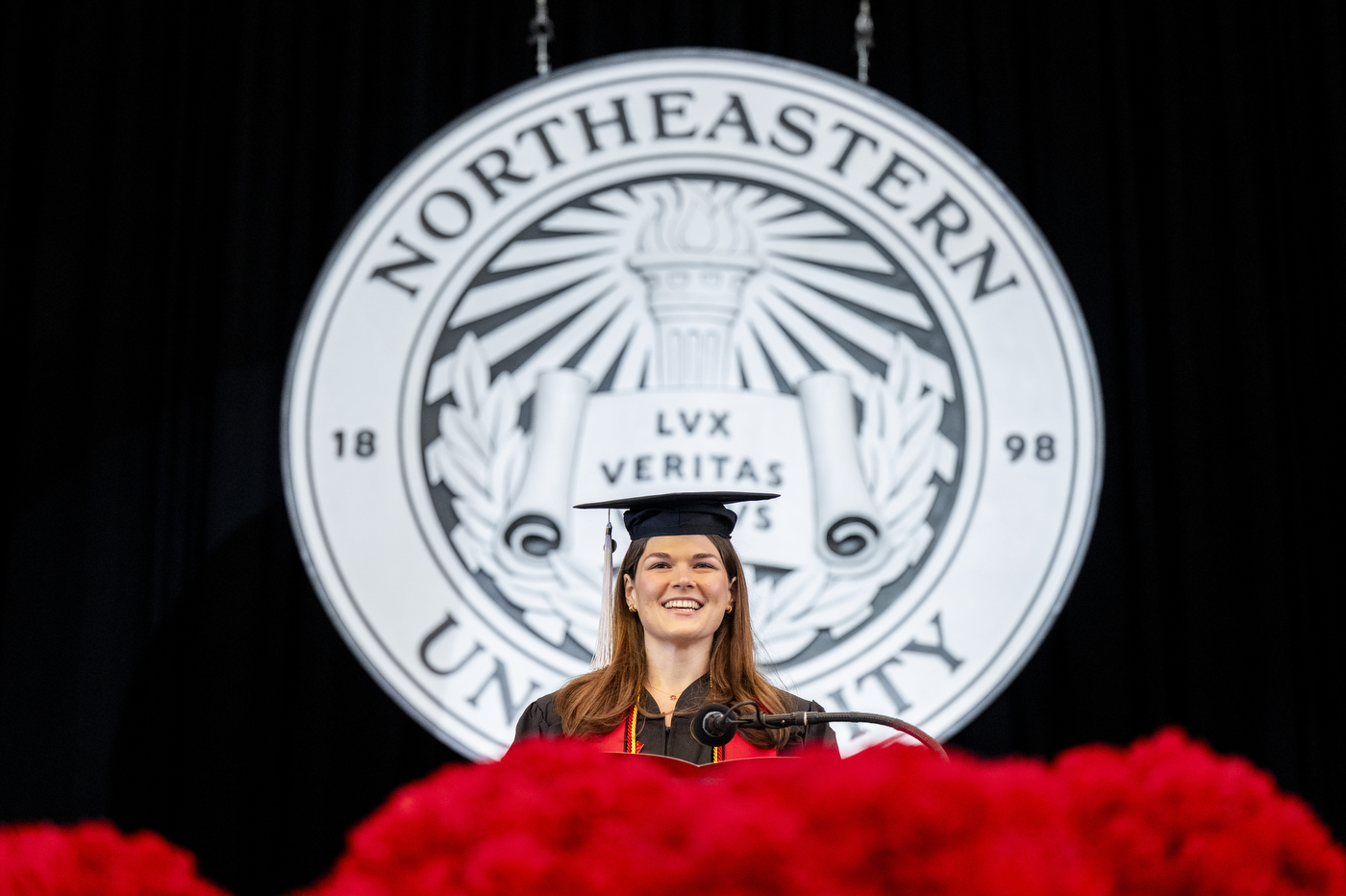 A graduate stands on stage in a cap, in front of the Northeastern logo.