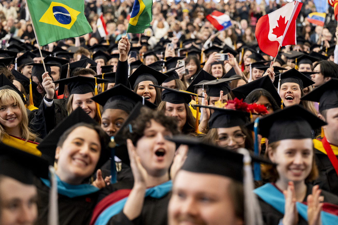 A crowd of graduates in caps and gowns, waving different country flags including Brazil and Canada.