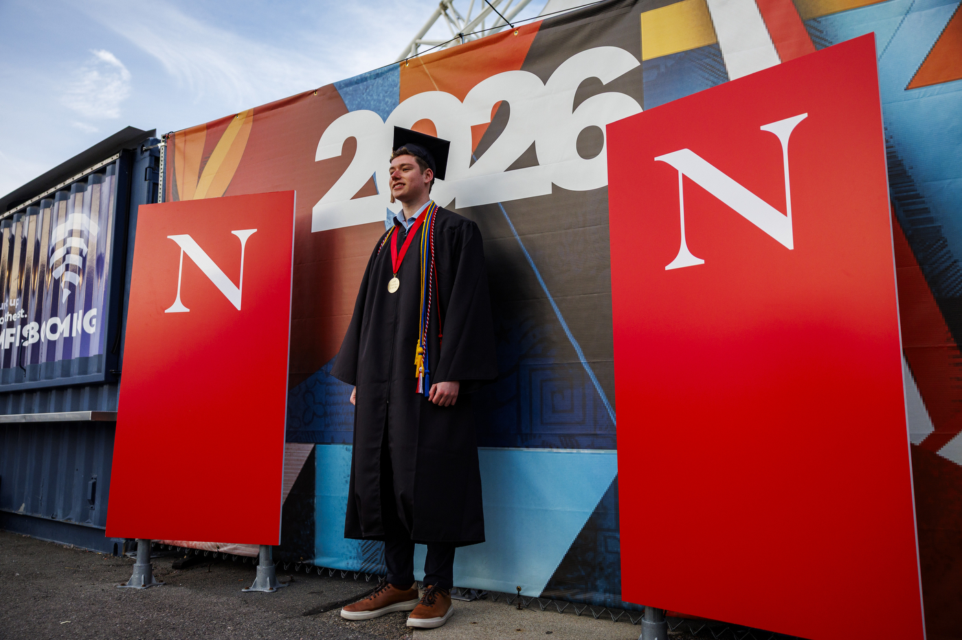 A student takes a photo at the D'Amore-McKim School of Business 2026 Undergraduate Commencement at Leader Bank Pavilion, with large red and blue '2026' and Northeastern 'N' banners visible on stage.