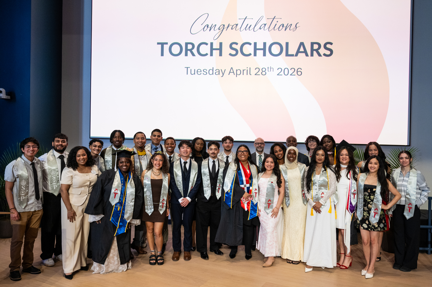 The Torch Scholars students standing on stage posing for a group photo with Ken Henderson in front of a screen that says 'Congratulations Torch Scholars Tuesday April 28th 2026'.