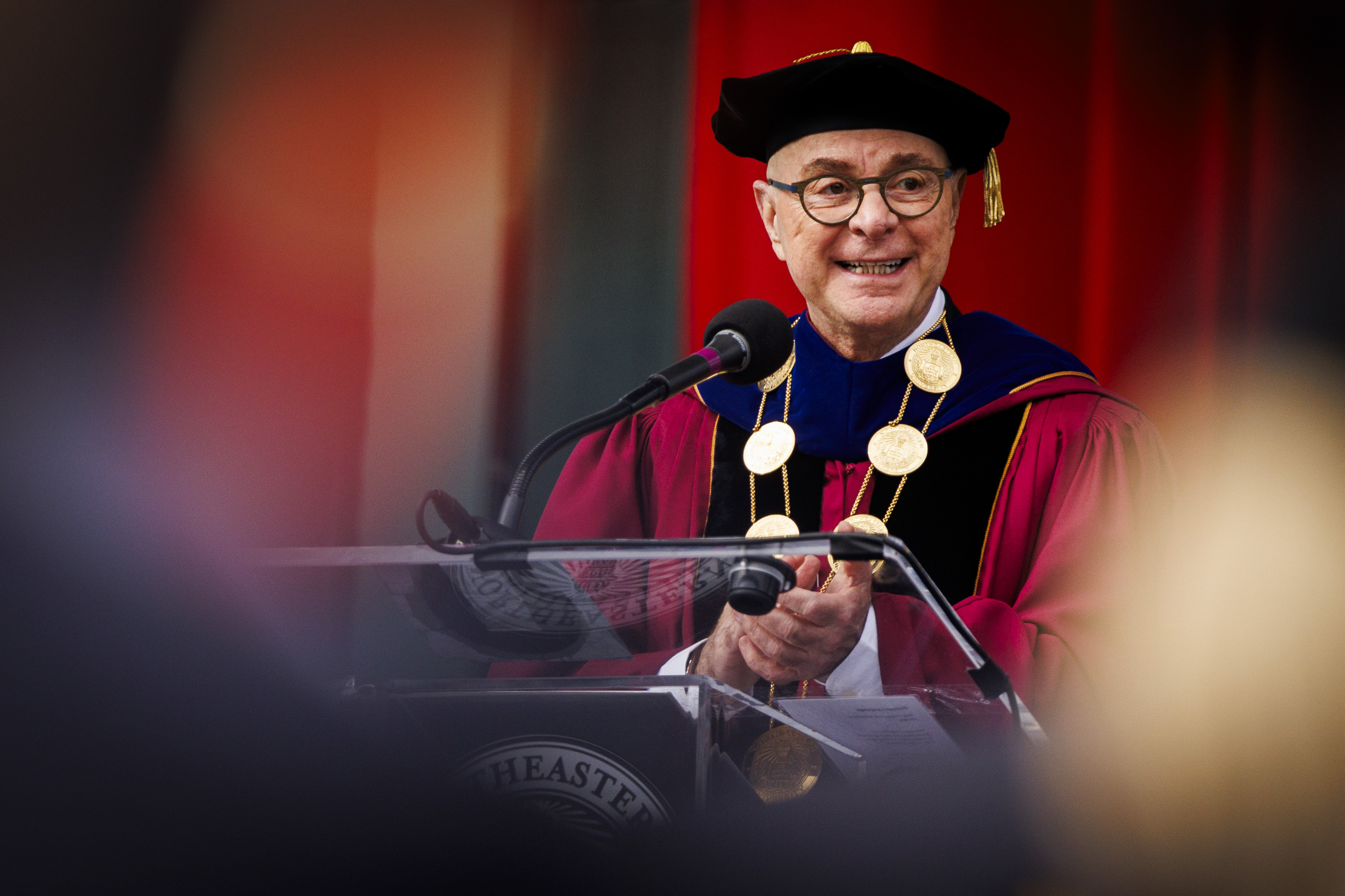 Northeastern University President Joseph E. Aoun speaks at a podium wearing crimson academic regalia with gold medallions during the 2026 commencement ceremony at Fenway Park.