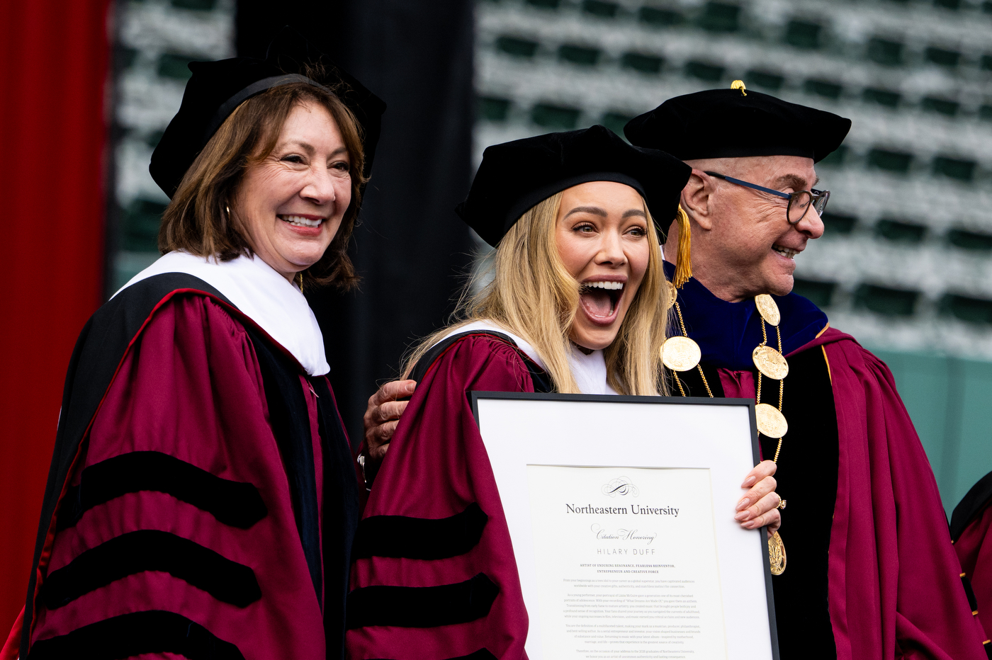 Hilary Duff, President Aoun, and Northeastern faculty member stand on stage. Duff holds an honorary degree and smiles wide.