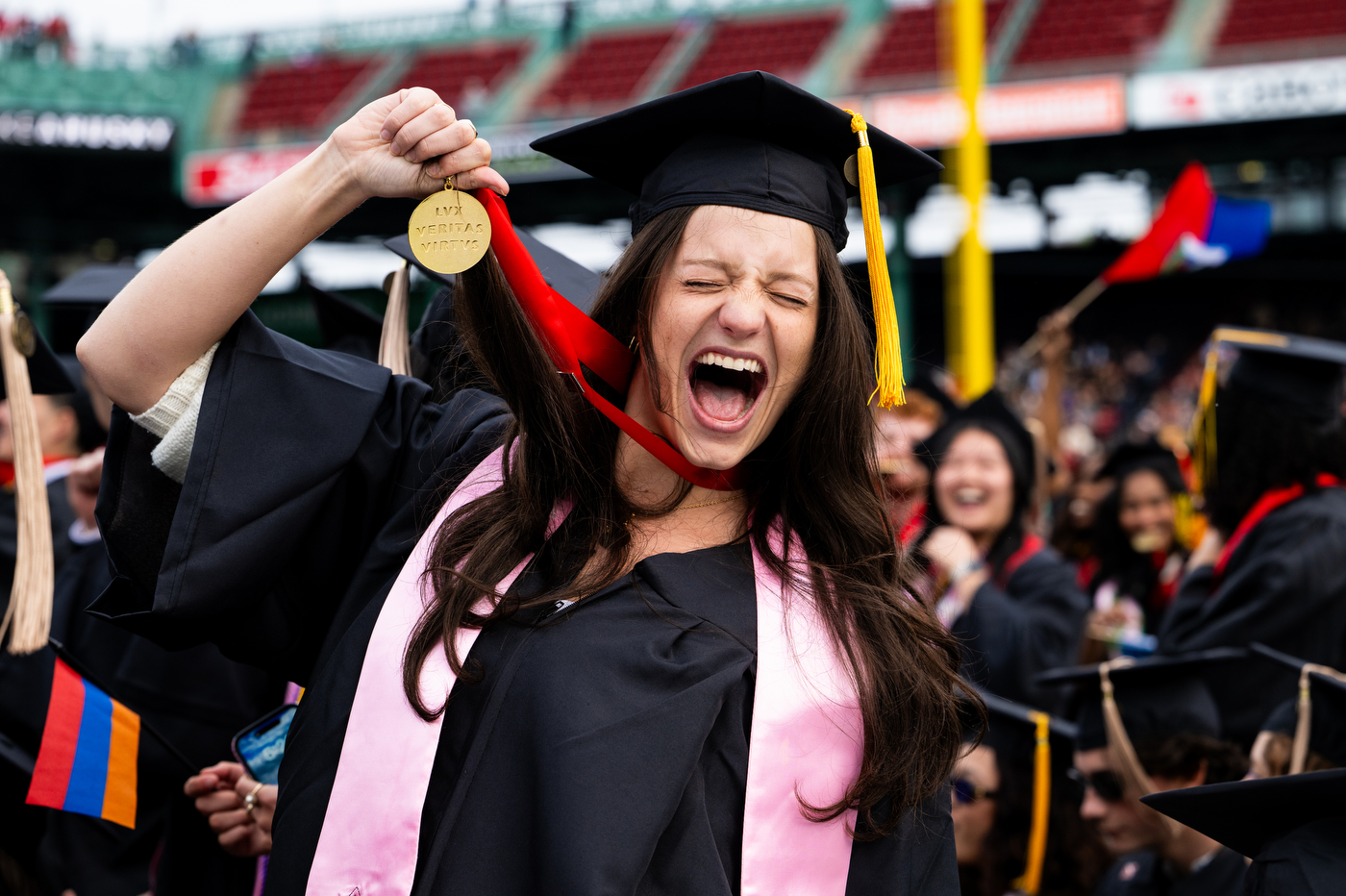 A graduating student celebrates holding up a Northeastern medal, with a crowd of other graduates in the background.
