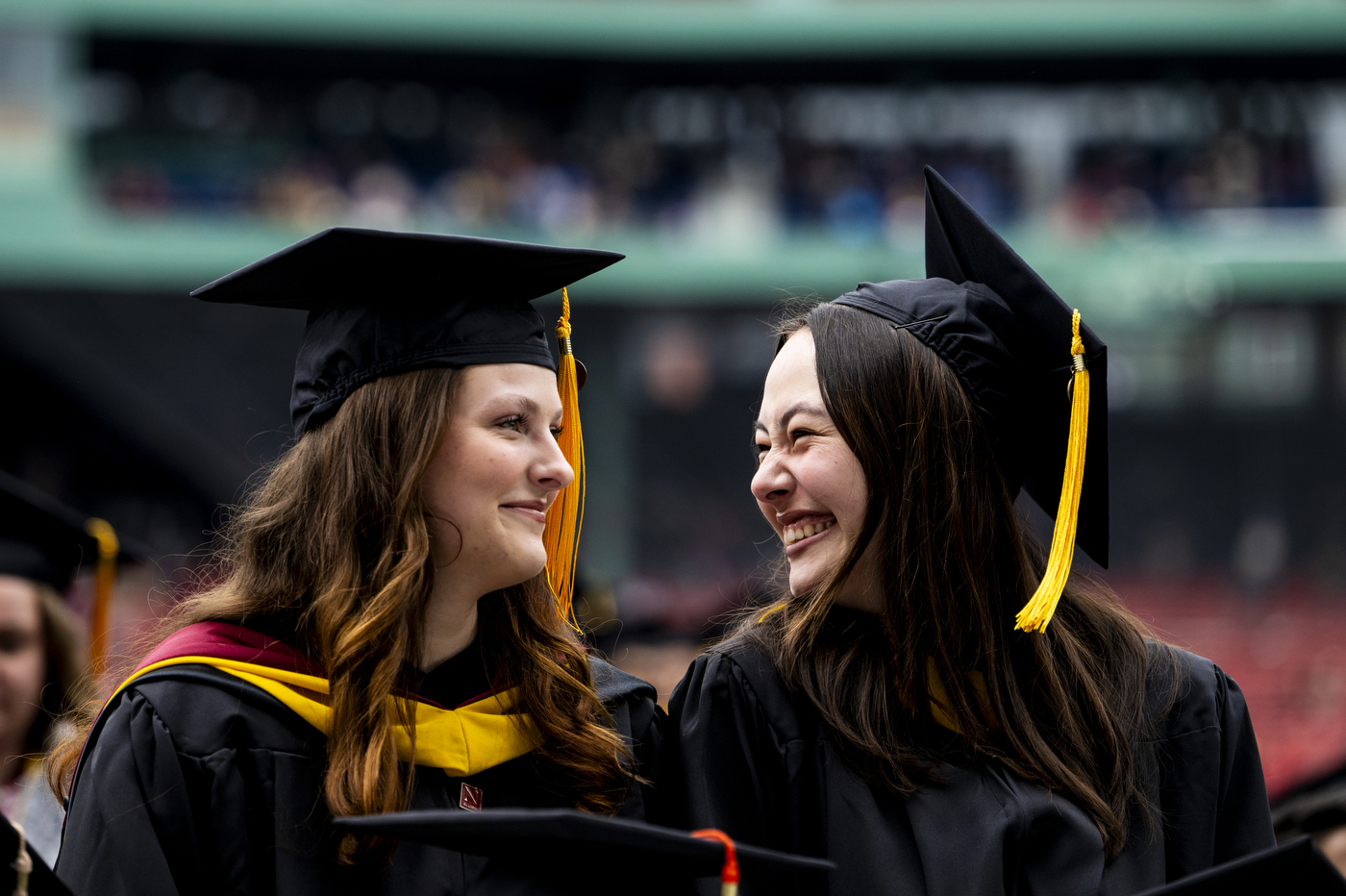 Two graduates wearing caps and gowns looking at each other and smiling in Fenway Park.