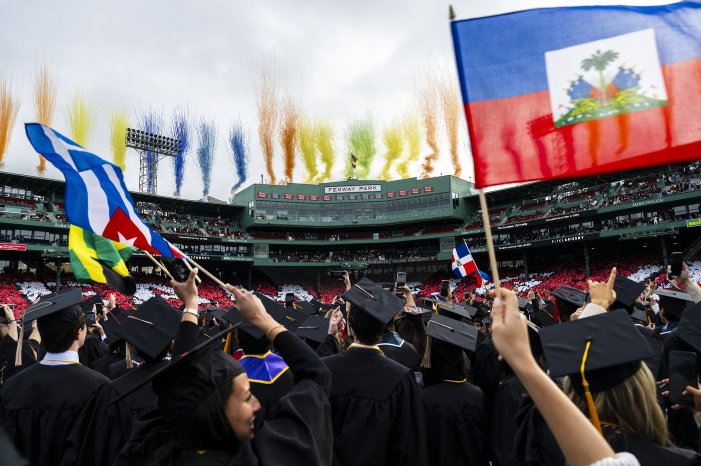 A wide shot of Fenway Park wtih colorfil fireowrks in the background while graduates cheer and wave country flags.