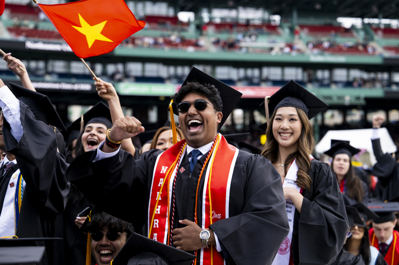 A jubilant Northeastern University graduate in sunglasses pumps his fist. Behind him someone waves a Vietnamese flag, surrounded by fellow graduates in caps and gowns.