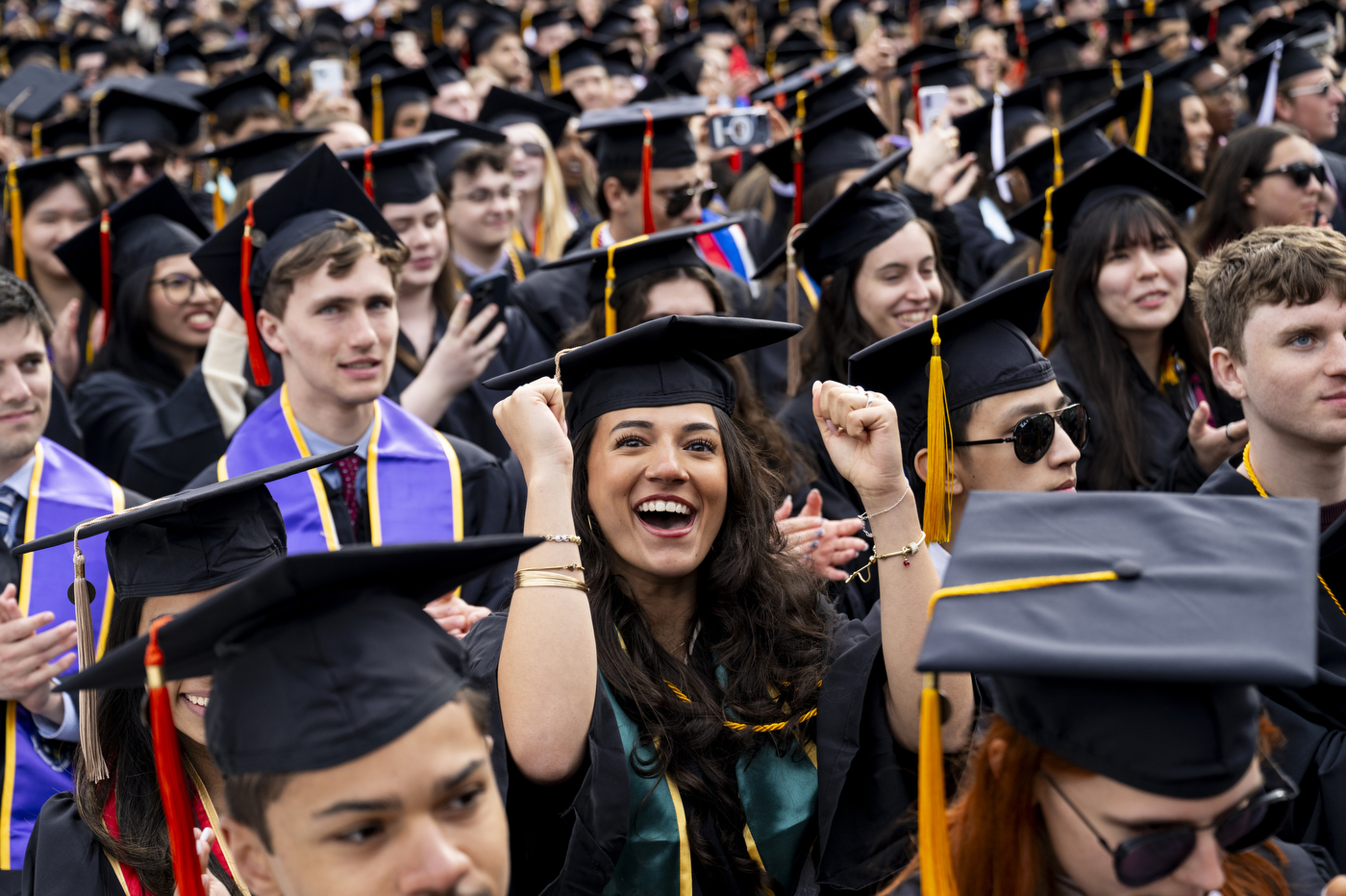 In a crowd of graduates facing the stage, on student smiles at the camera and pumps her fists.