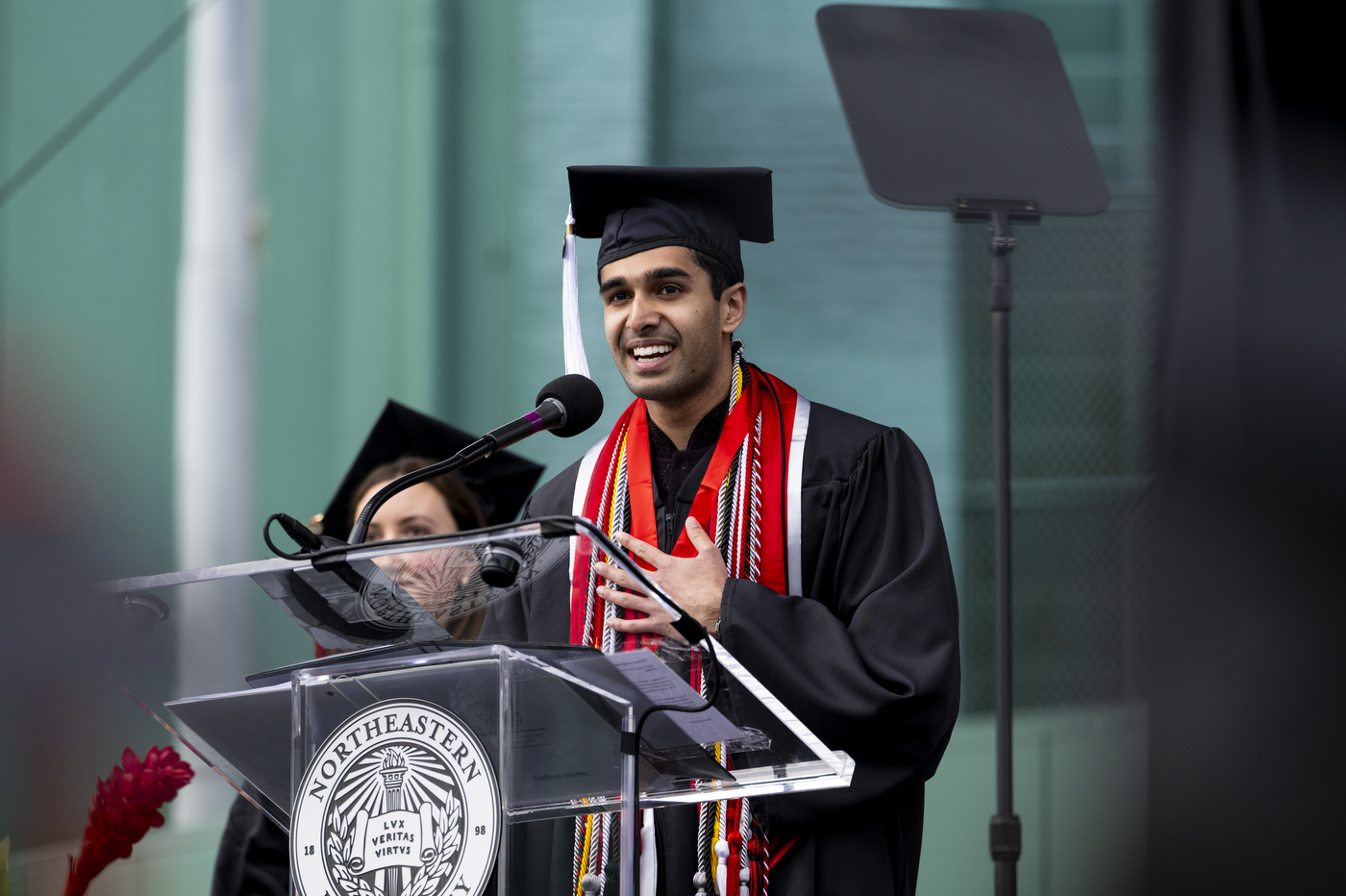 A student speaker stands at a podium wearing a cap and gown with one hand on their chest. 