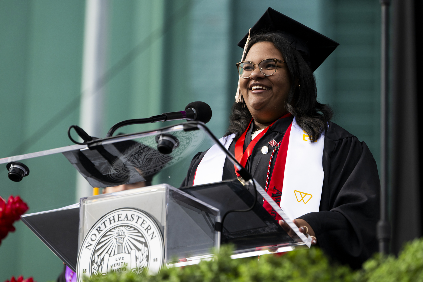 A student speaker wearing a cap and gown stands in front of a microphone smiling. 