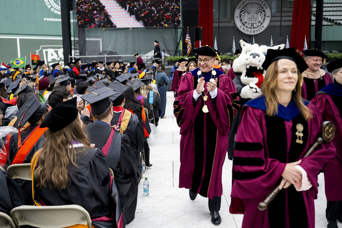 The academic procession enters Fenway Park for Northeastern University's 2026 undergraduate commencement, with the Northeastern Husky mascot visible among faculty in colorful regalia.