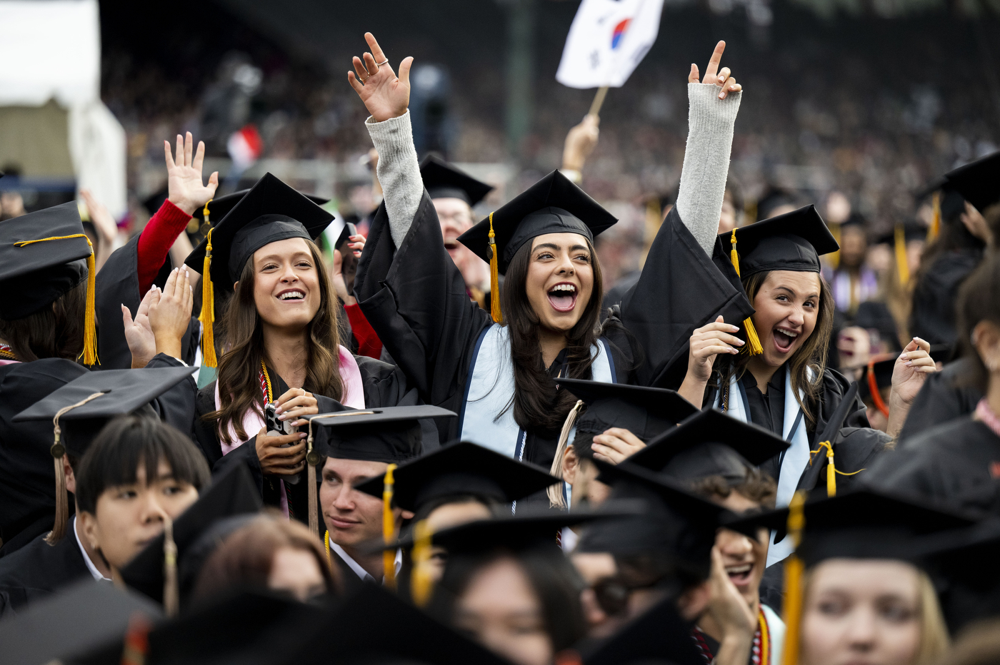 A group of Northeastern University graduates in black caps and gowns laugh and raise their hands joyfully during the 2026 commencement ceremony at Fenway Park, with a South Korean flag visible in the background.