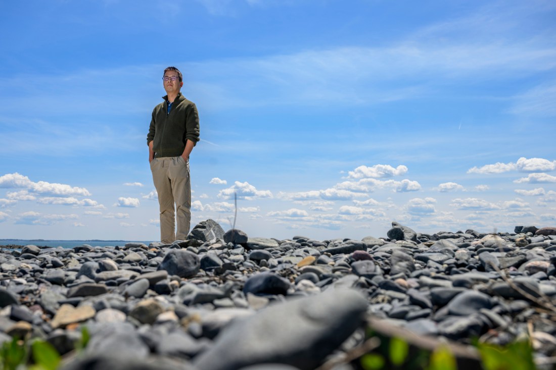 Portrait of Chengfei He standing on a rocky beach in Nahant with blue sky behind him. 