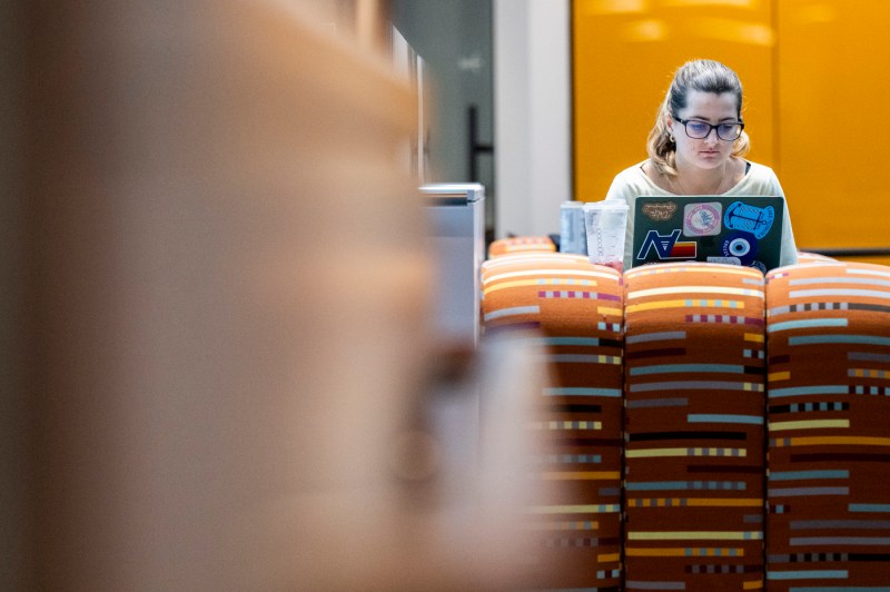 A shot of a student using a laptop while sitting on an orange chair, with a blur effect on the left side of the image.