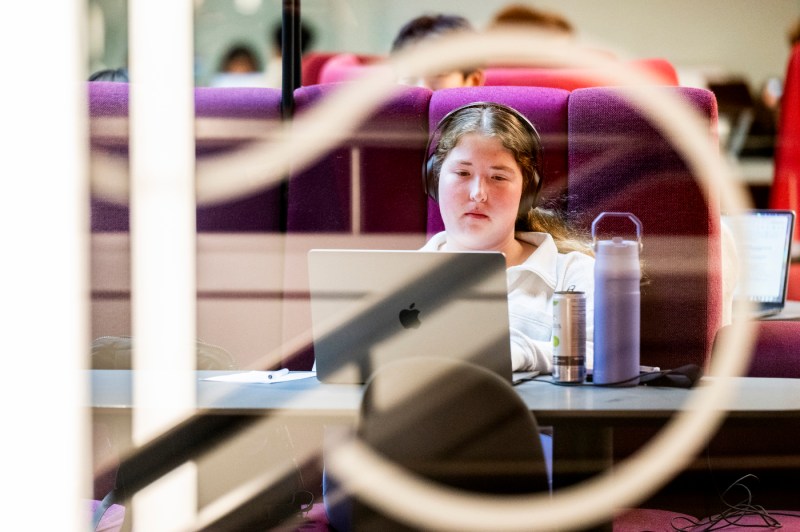 A close up of a student wearing headphones and using a laptop while sitting on a purple seat.