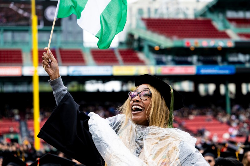 A joyful Northeastern University graduate in a black cap and gown waves a green and white Nigerian flag at Fenway Park during the 2026 commencement ceremony, laughing with excitement.
