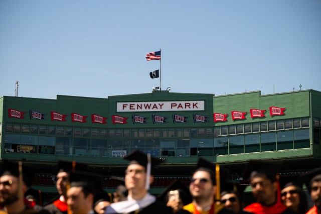 Shows graduates at the stadium with Fenway Park sign and flags in the background.
