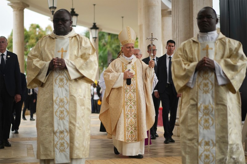 Pope Leo XIV, dressed in gold and white papal vestments and mitre, carries a ceremonial staff while walking in a procession flanked by two priests in matching robes.