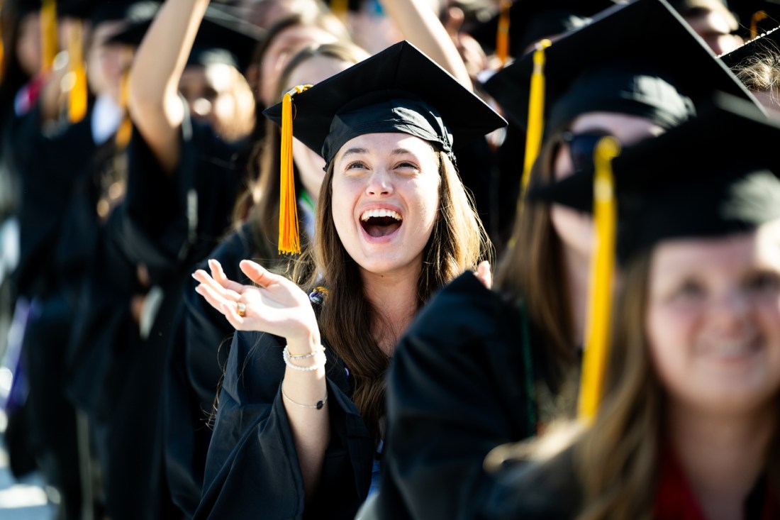 A person wearing a cap and gown cheers and claps excitedly at the 2025 undergraduate Commencement ceremony at Fenway park.