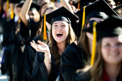 A person wearing a cap and gown cheers and claps excitedly at the 2025 undergraduate Commencement ceremony at Fenway park.