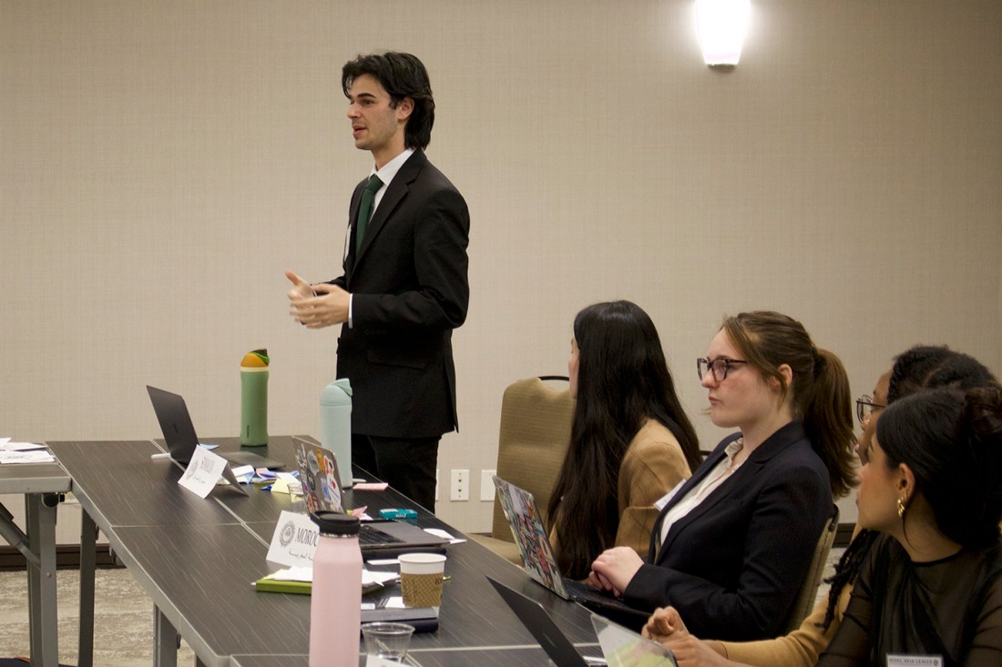 Student delegate in a suit speaks while others listen at a committee table with laptops and notes.