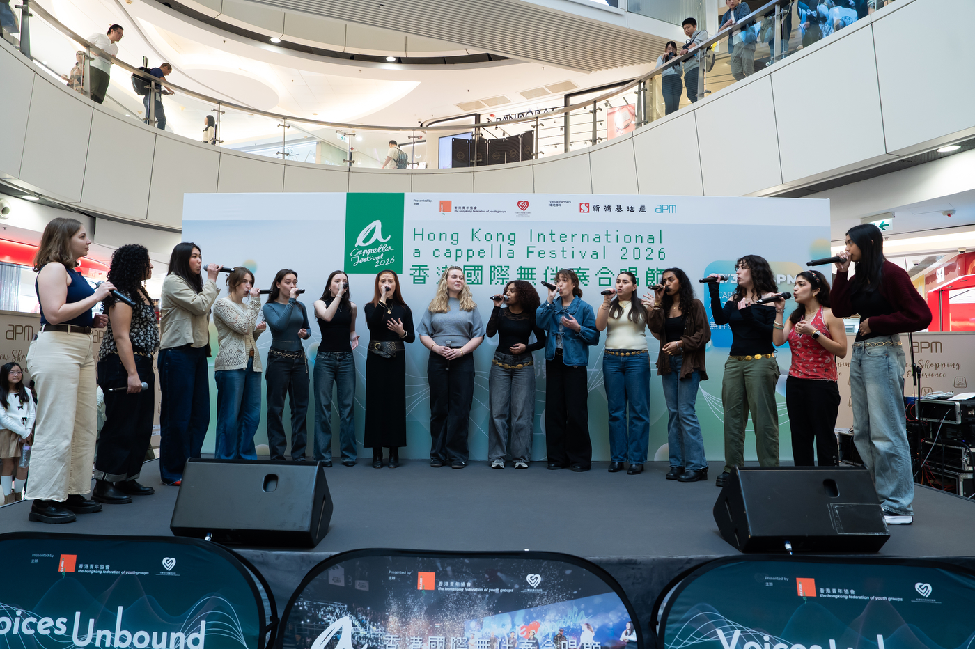 Members of Pitch, Please! perform on an indoor stage at the Hong Kong International A Cappella Festival, standing in a line in front of large speakers in a shopping mall atrium. 