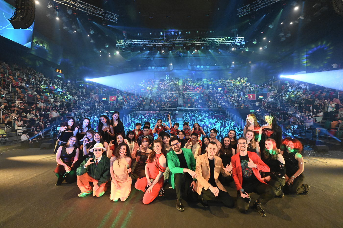 A group of a cappella performers poses together on stage for a group photo with a packed crowd and stage lighting visible behind them. 