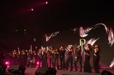 A group of a capella performers dressed in black stand on a dimly lit stage in an indoor event, arms raised and gesturing while they sing into microphones.