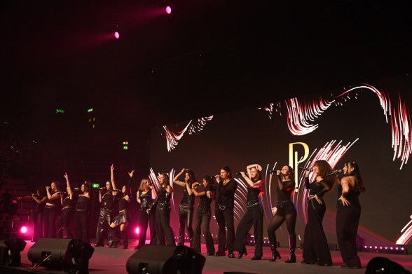 A group of a capella performers dressed in black stand on a dimly lit stage in an indoor event, arms raised and gesturing while they sing into microphones.