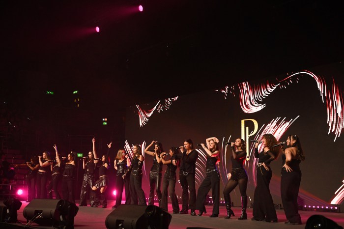 A group of a capella performers dressed in black stand on a dimly lit stage in an indoor event, arms raised and gesturing while they sing into microphones.