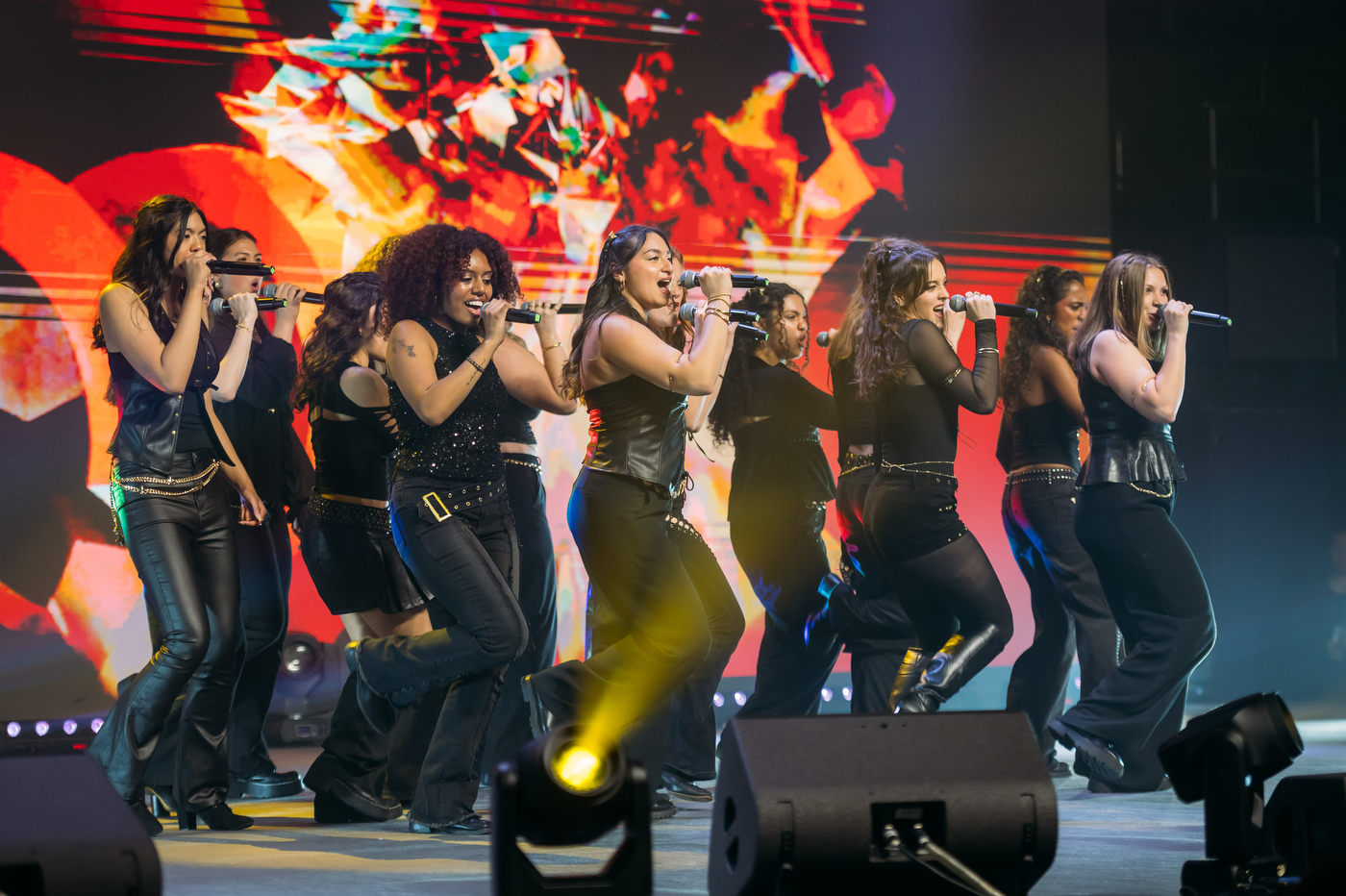 A women's a cappella group performs with energy on a concert stage, singing into microphones with a colorful LED backdrop behind them. 