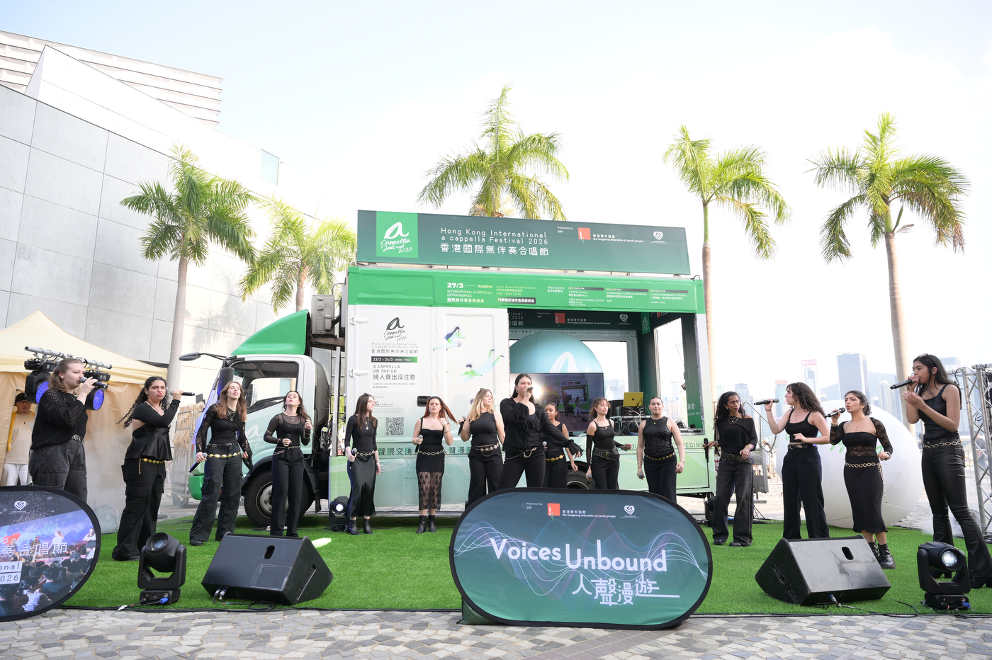 A student a cappella group wearing all black performs outdoors on a turf stage with "Voices Unbound" signage in front and palm trees behind them. 