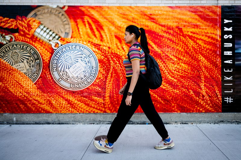 A students walks in front of a bright orange mural showcasing Northeastern medallions. 