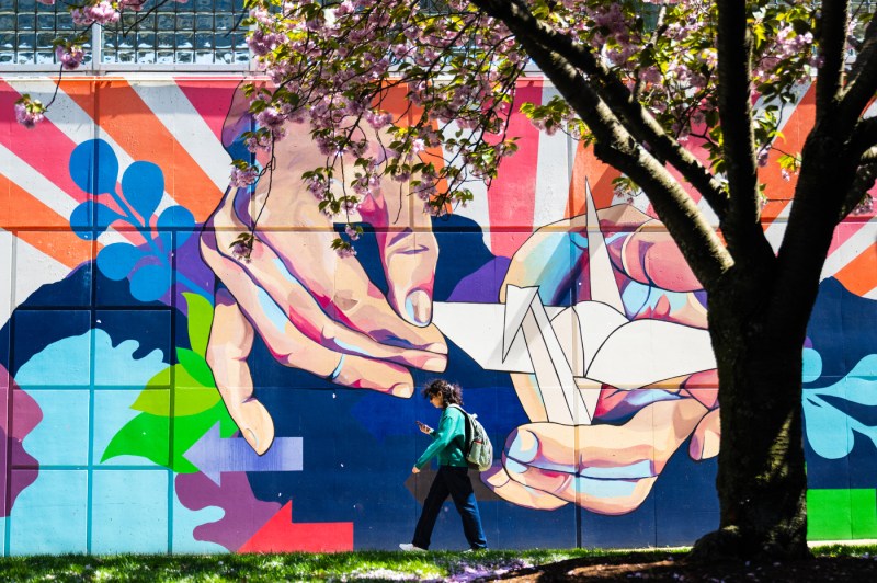A students walks in front of a bright colorful mural showcasing two hands folding a paper crane.
