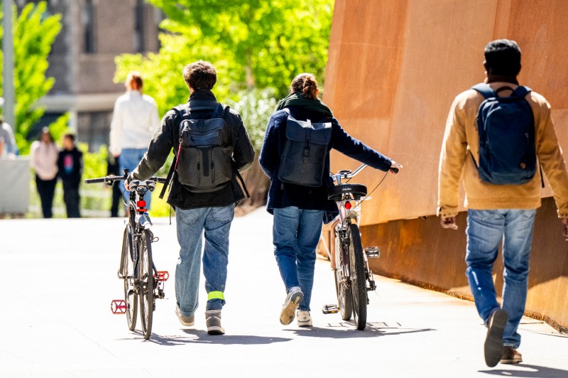 Three people walk over the pedestrian bridge. Two of them wear backpacks and walk with their bikes.
