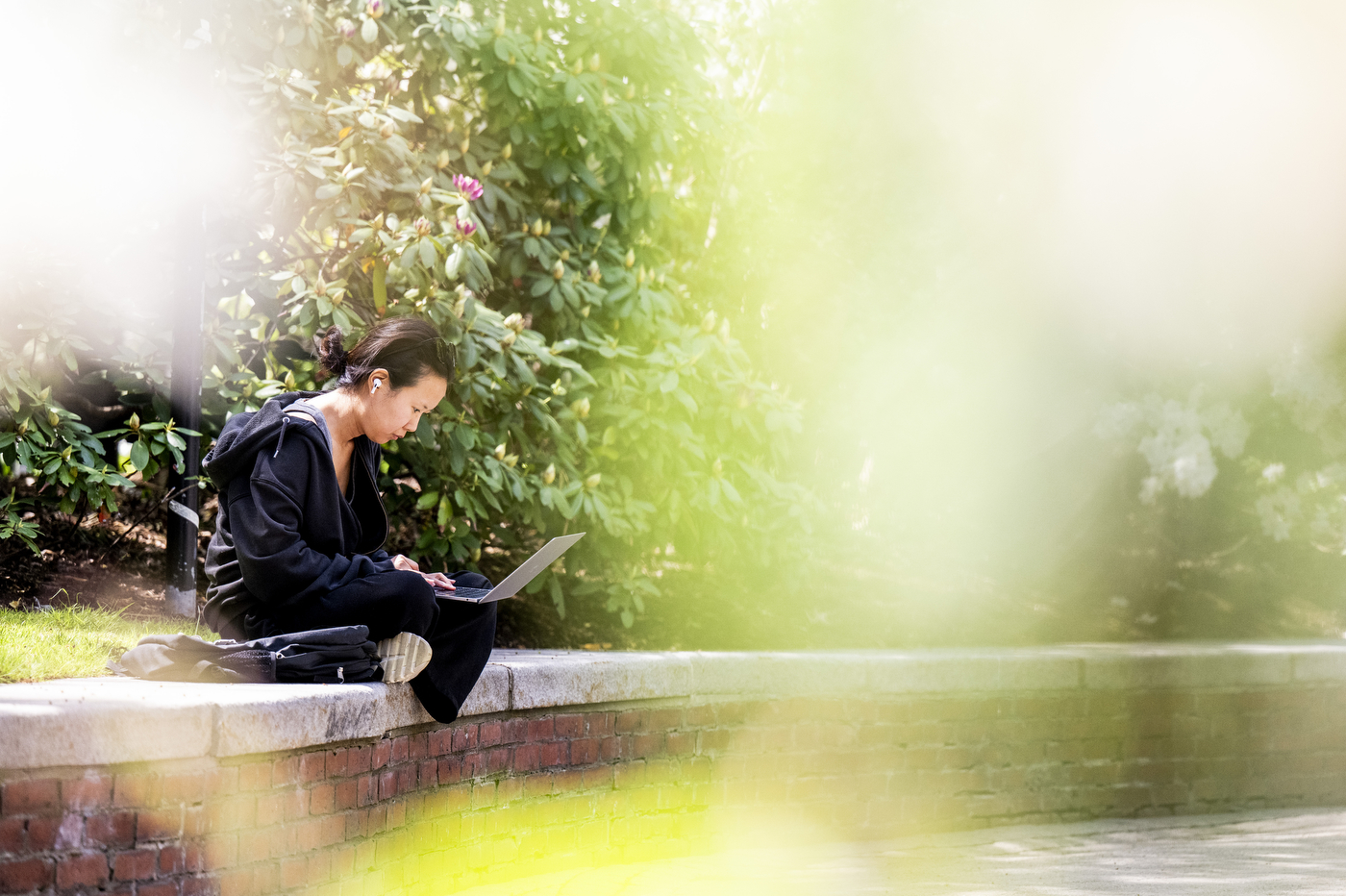 A students sits outside on a low brick wall while working on a laptop.